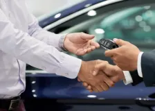 Two professionals shake hands while exchanging a car key fob beside a vehicle, symbolizing a vehicle sale, lease agreement, or dealership transaction.