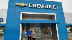 Two men and one woman standing in front of blue walled Chevrolet dealership entrance.
