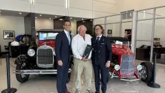 Three men standing in front of two classic cars handing off gift. 