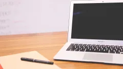 Laptop computer on a desk with a notepad and pen next to it and a whiteboard in the background