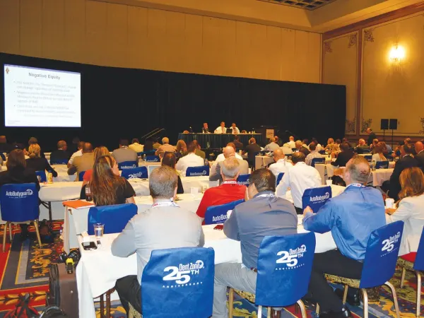 Audience seated at tables attending an automotive industry conference session, with a panel of speakers presenting on stage and a presentation slide displayed on a large screen.