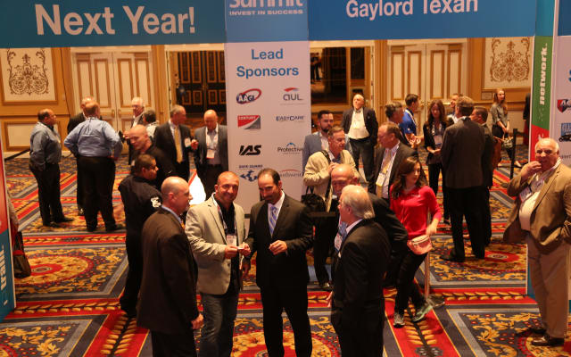  Attendees networking and talking in a conference hall beneath signage for the Industry Summit with sponsor displays in the background.