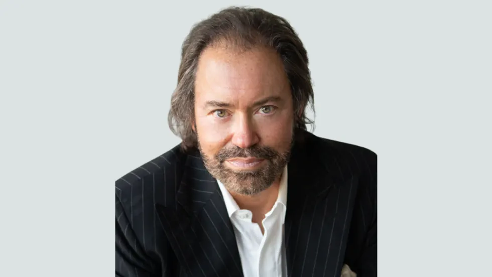 A man in a pinstripe suit jacket and white collared shirt looks directly at the camera with a neutral expression, seated against a light gray background.
