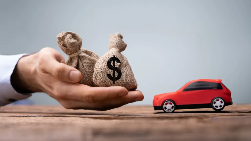 A hand holding small burlap money bags next to a toy red car, symbolizing auto financing, loan payments, and dealership profitability.
