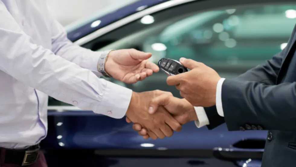 Two professionals shake hands while exchanging a car key fob beside a vehicle, symbolizing a vehicle sale, lease agreement, or dealership transaction.