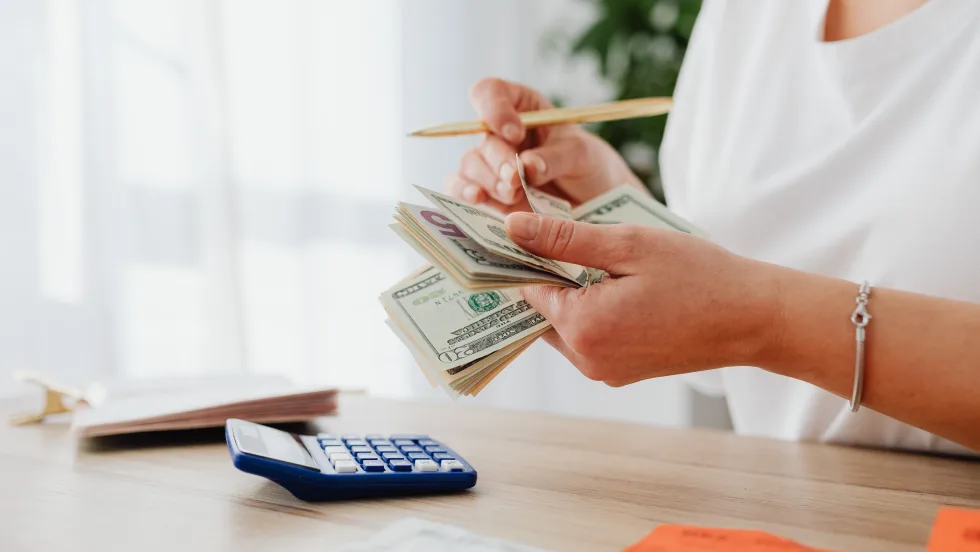 Photo of woman counting cash with a calculator sitting on a desk in front of her