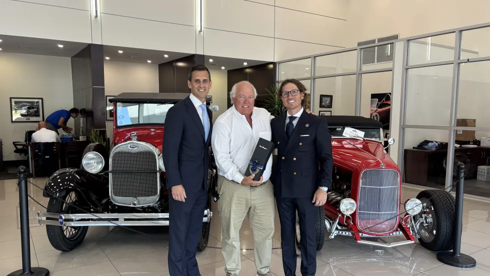 Three men standing in front of two classic cars handing off gift. 