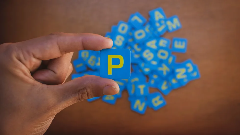 Photo of man's hand holding a square blue piece with a yellow capital P on it, with other letter pieces below on a surface