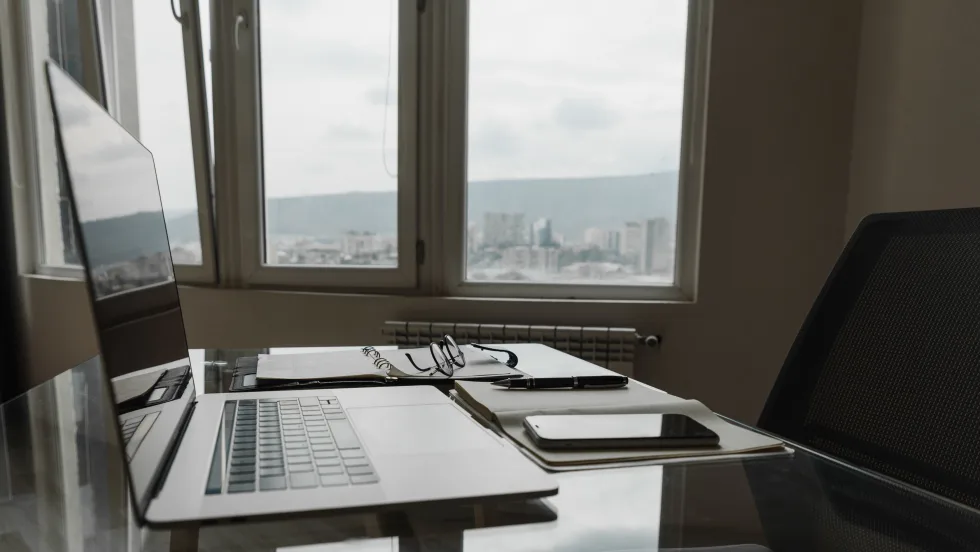 Photo of a laptop, cellphone and eyeglasses on a desk next to an office window