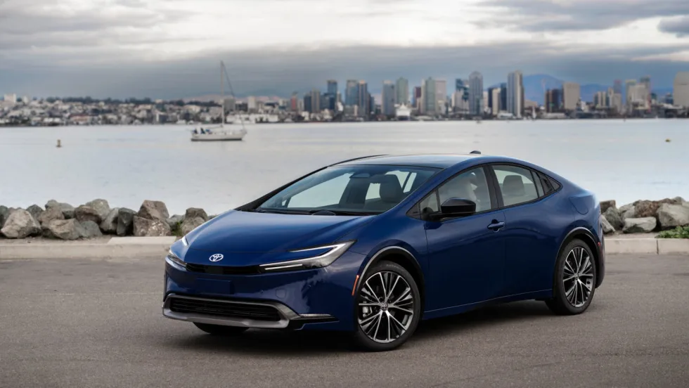 Photo of blue Toyota Prius parked next to a body of water with a city skyline behind it