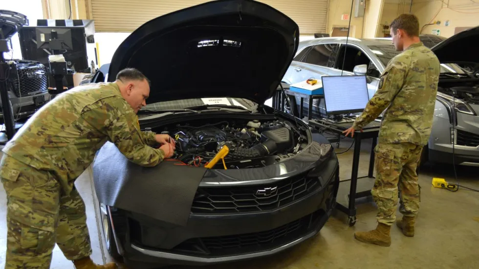 two men in Army uniforms looking under the hood of a car