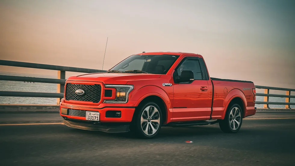 red pickup truck in front of railing and body of water with setting sun