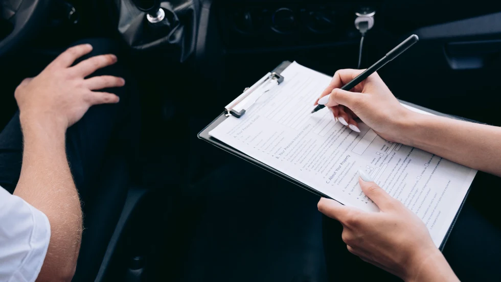two people sitting in a car, one in the driver seat, one in the passenger seat with a clipboard and pen
