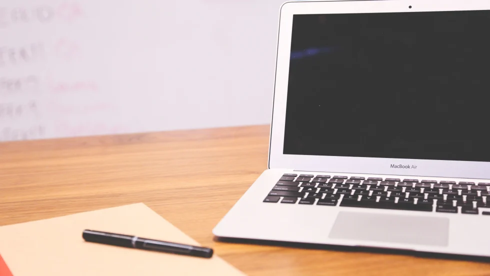 Laptop computer on a desk with a notepad and pen next to it and a whiteboard in the background