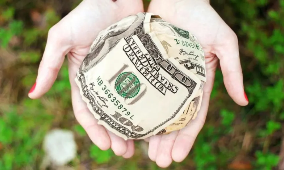 Hands holding a ball made of folded U.S. hundred-dollar bills, symbolizing financial assistance or giving.