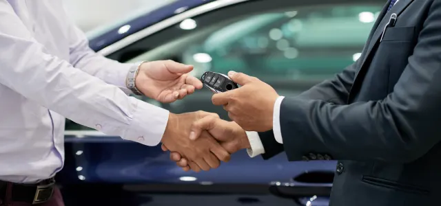 Two professionals shake hands while exchanging a car key fob beside a vehicle, symbolizing a vehicle sale, lease agreement, or dealership transaction.