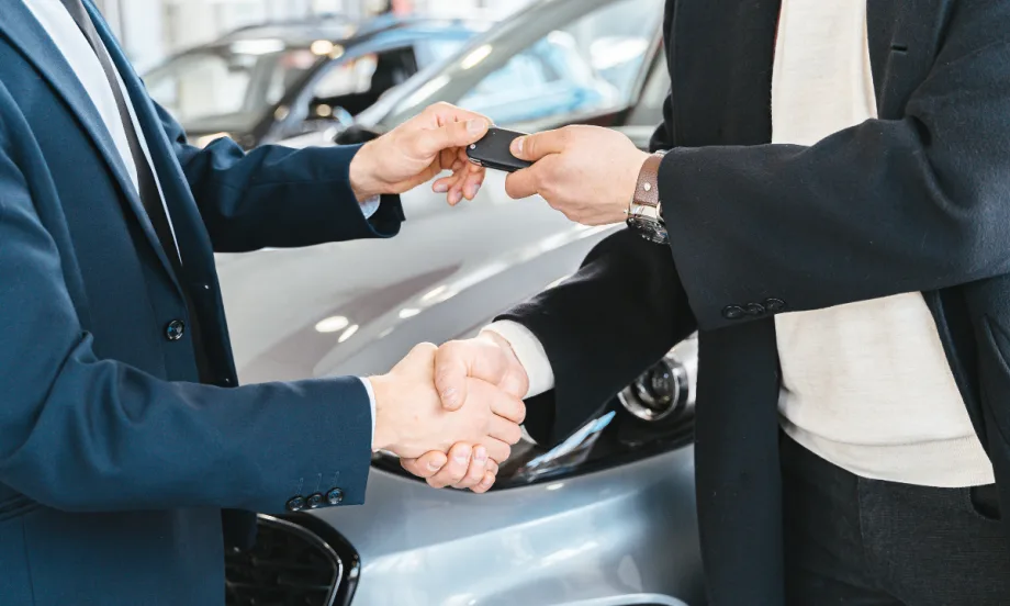 two men shaking hands with one handing over a set of car keys, a car in the background