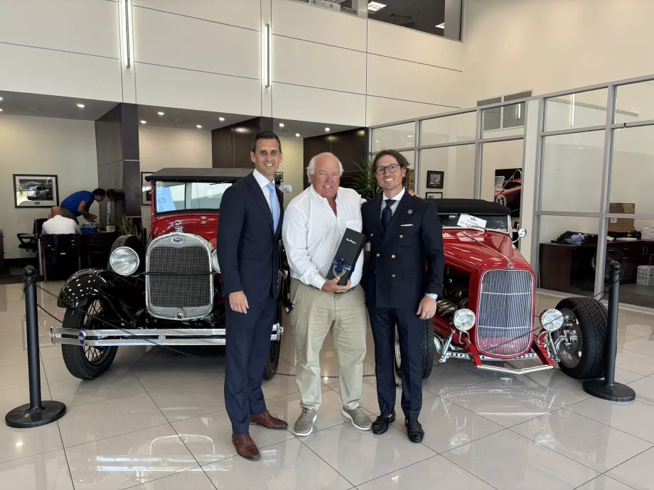 Three men standing in front of two classic cars handing off gift.