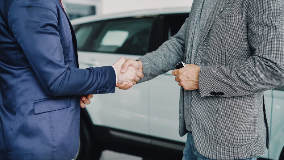 Two men in suit jackets shaking hands in front of a new-looking white vehicle