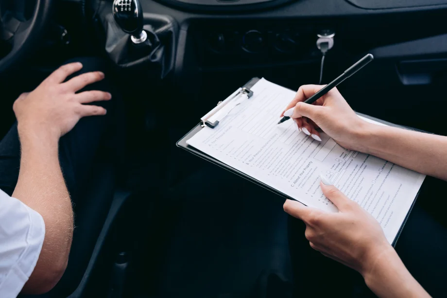 two people sitting in a car, one in the driver seat, one in the passenger seat with a clipboard and pen