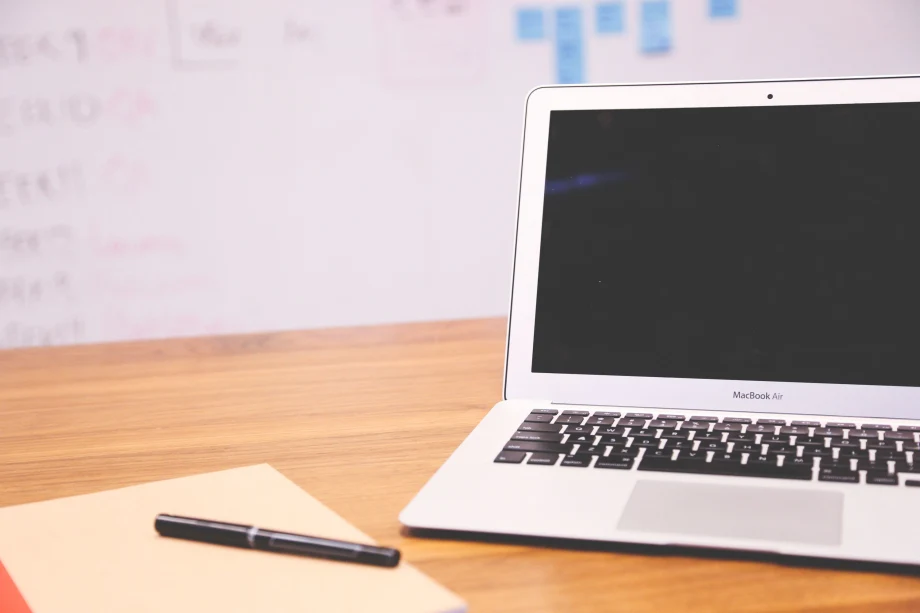 Laptop computer on a desk with a notepad and pen next to it and a whiteboard in the background